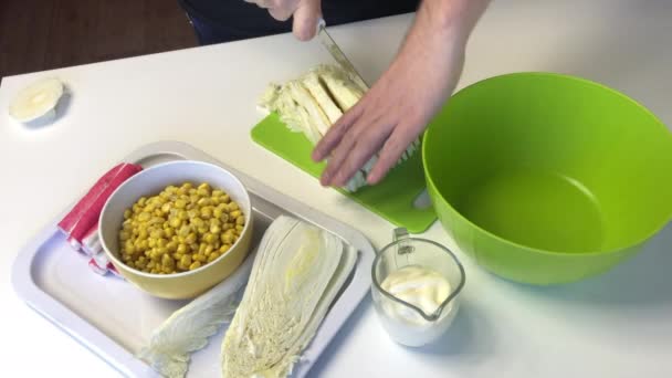 Een man maakt een salade van Beijing kool. Snijdt kool met een mes op een snijplank. Naast de tafel liggen nog andere ingrediënten. Maïs, krab en mayonaise — Stockvideo