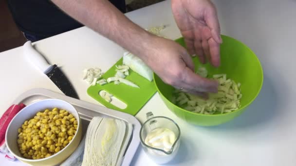 Человек делает салат из пекинской капусты. Нарезает капусту ножом на разделочной доске и бросает в контейнер. Рядом со столом есть и другие ингредиенты. Кукуруза, крабовые палочки и майонез . — стоковое видео