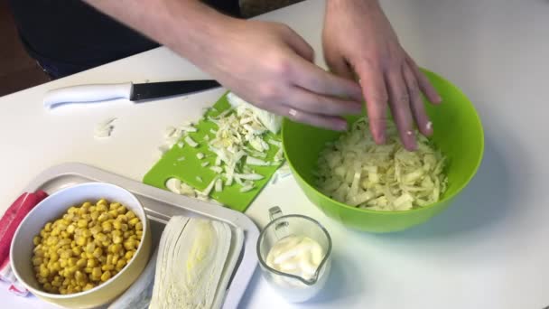 En man gör en sallad av Pekingkål. Skivor kål med en kniv på en skärbräda och kastar den i en behållare. Bredvid bordet finns andra ingredienser. Majs, krabbpinnar och majonnäs — Stockvideo