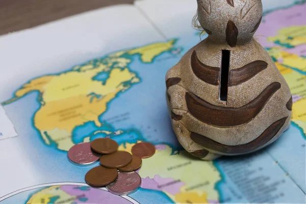 A ceramic piggy bank with a slot for coins is on the world map. Several coins are scattered nearby. Piggy bank for travel.
