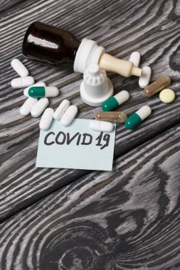 Üzerinde Covid-19 yazan kağıt çıkartması. Yakınlarda çeşitli şekil ve renklerde birkaç hap var. Burun spreyli kavanozlar. Fırçalanmış çam ağaçlarının üzerinde.