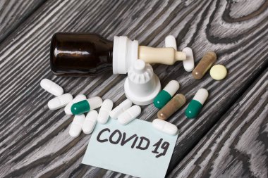 Üzerinde Covid-19 yazan kağıt çıkartması. Yakınlarda çeşitli şekil ve renklerde birkaç hap var. Burun spreyli kavanozlar. Fırçalanmış çam ağaçlarının üzerinde.