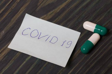 Üzerinde Covid-19 yazan kağıt çıkartması. Yakınlarda beyaz-yeşil renkli birkaç hap var. Fırçalanmış çam ağaçlarının üzerinde.