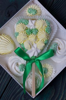 Çubukta kalp şeklinde şekerlemeler. Beyaz, sarı ve yeşil elementlerden yapılmış. Fiyonkla süslenmiş. Karton bir kutuda yatıyor. Onun yanında farklı şekilli şekerlemeler var..