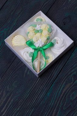 Çubukta kalp şeklinde şekerlemeler. Beyaz, sarı ve yeşil elementlerden yapılmış. Fiyonkla süslenmiş. Karton bir kutuda yatıyor. Onun yanında farklı şekilli şekerlemeler var..