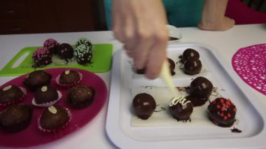 Kadın çikolatalı sünger toplarını damla damla çikolatayla süslüyor. Patates keki pişiriyorum. Tabakların yanında kek kurusıkı, sıvı çikolata ve renkli serpinti var..