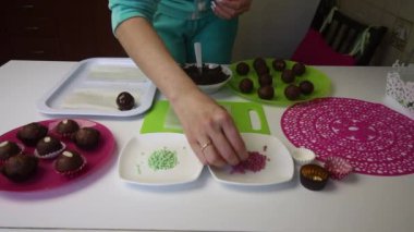 Bir kadın çikolatalı topları sünger kekine serpiyor. Patates keki yapıyor. Tabakların yanında kek kurusıkı, sıvı çikolata ve renkli serpinti var..