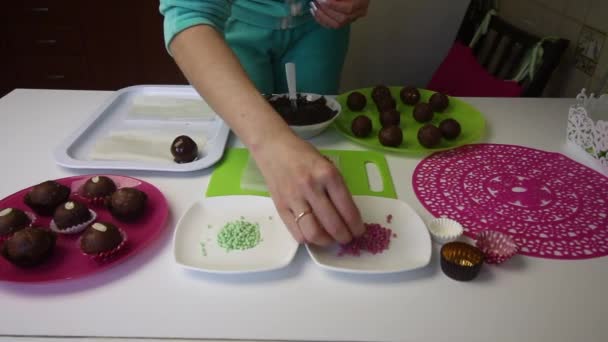 Una mujer espolvorea bolas de chocolate en un bizcocho. Hace un pastel de patata. Junto a los platos hay pasteles en blanco, chocolate líquido y chispas de colores. . — Vídeos de Stock