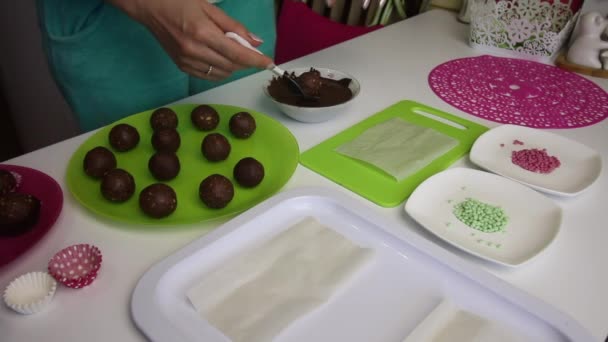 Жінка занурює кульки з губного торта в шоколад. Робить картопляний торт. Поруч з тарілками є заготовки для торта, рідкий шоколад і кольорові зморшки . — стокове відео