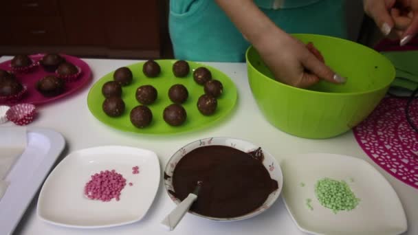 Woman rolls balls of biscuit mass with her hands. The mass is brown, made from crumbs of cookies and cocoa, with the addition of butter. Next to the plates are cake blanks, liquid chocolate and colore — Stock Video