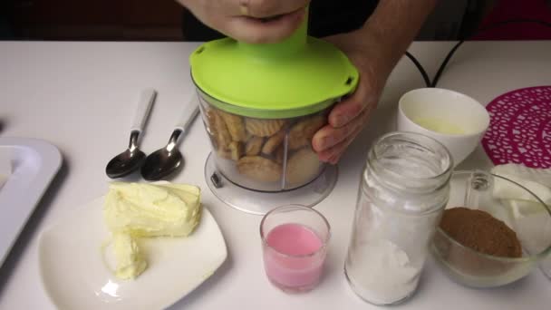 Cookies hälls i mixerskålen. En man kopplar ihop en apparat och krossar kakor i en skål. Övriga ingredienser för att göra Potato cookies anges i närheten. — Stockvideo