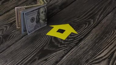 Fırçalanmış tahtaların üzerinde dolar banknotları var. Yakınlarda kağıttan kesilmiş bir evin silueti var. Adamın biri üzerine bir sürü anahtar koymuş..