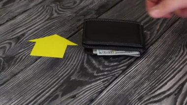 Fırçalanmış tahtalardan oluşan bir masanın üzerinde kağıttan kesilmiş bir evin silueti yatıyor. Adamın biri üzerine cüzdan koymuş. Dolarlar buradan görülebiliyor. Ondan sonra, adam üzerine bir sürü anahtar koydu..