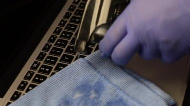 Lastik eldivenli bir adam laptopunu temizliyor. Bir sprey şişesinden mendile deterjan sürüyor ve dizüstü bilgisayarın klavyesini siliyor. Çalışan ekipmanların viral enfeksiyondan korunması.