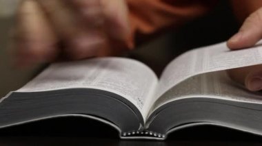 Masanın üzerinde açık bir İncil yatıyor. Adam arzulanan bölümü bulmak için sayfaları hızla çeviriyor. Okuyor, çizgiler boyunca bir parmağını hareket ettiriyor. Yakın çekim.