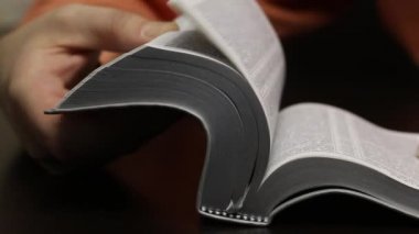 Masanın üzerinde açık bir İncil yatıyor. Adam arzulanan bölümü bulmak için sayfaları hızla çeviriyor. Okuyor, çizgiler boyunca bir parmağını hareket ettiriyor. Yakın çekim...