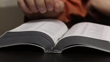 Masanın üzerinde açık bir İncil yatıyor. Bir adam okuyor, parmağını çizgiler boyunca hareket ettiriyor, sayfaları çeviriyor. Yakın çekim.
