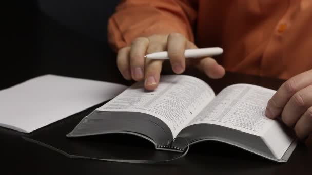 Человек читает Библию, сидя за столом. Указывает пальцем по линии. Пишет важные мысли на листе бумаги. Крупный план — стоковое видео