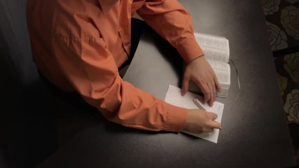 Um homem lê a Bíblia enquanto está sentado à mesa. Escreve pensamentos importantes em um pedaço de papel. Corrige um marcador e fecha o livro . — Vídeo de Stock