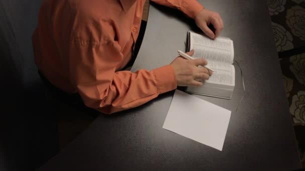 Um homem lê a Bíblia enquanto está sentado à mesa. Leva um dedo ao longo das linhas. Escreve pensamentos importantes em um pedaço de papel . — Vídeo de Stock