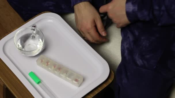 Ένας άνθρωπος παίρνει ένα θερμόμετρο υδραργύρου από τη μασχάλη του, κοιτάζει την κατάθεσή του και την αποτινάζει. Μπροστά του σε ένα δίσκο είναι ένα κουτί δισκίων για πολλά κύτταρα και ένα ποτήρι νερό.. — Αρχείο Βίντεο