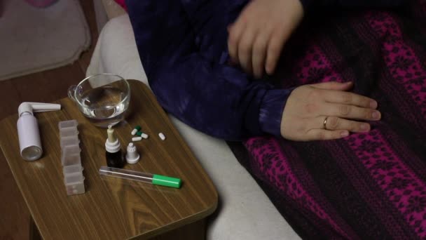 El enfermo yace en una cama cubierta con una manta. Toma un termómetro de mercurio de su axila y lo pone sobre una mesa. Junto a la silla hay varios medicamentos . — Vídeos de Stock