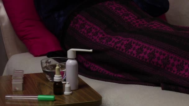 O homem doente está deitado numa cama coberta com um cobertor. Ele pega um termômetro de mercúrio de sua axila, balança-o e coloca-o em um suporte. Ao lado da cadeira estão vários medicamentos . — Vídeo de Stock