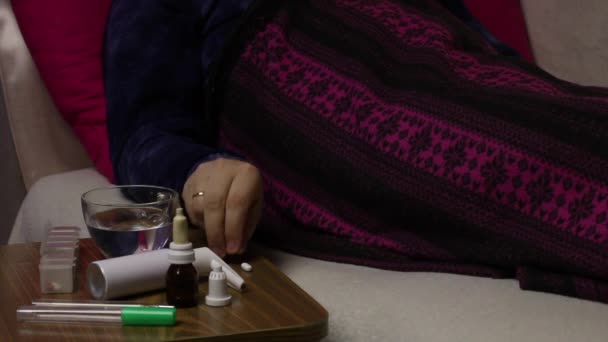 The sick man lies in a bed covered with a blanket. He takes a pill and drinks it with water. Next to the chair are various medications. — Stock Video