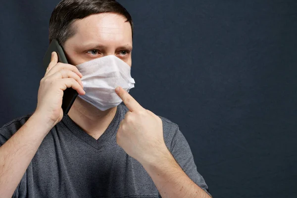 A man in a medical mask is talking on a smartphone. During a conversation does shows a finger on the mask. Protection against infection in self-isolation during coronavirus.