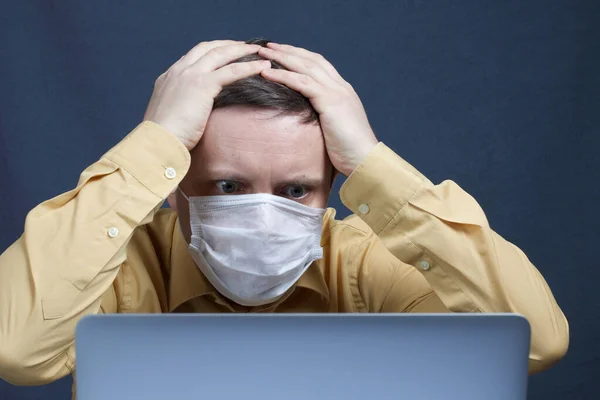医療マスクの男がノートパソコンでニュースを見ている ショックを受けて両手で頭を出す コロナウイルスの自己分離時の感染からの保護 — ストック写真