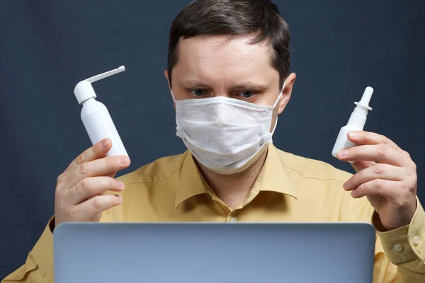 A man in a medical mask communicates via video conferencing on a laptop. In one hand he holds a nasal spray, in the other a spray for the throat and shows them to the interlocutor. Protection against infection in self-isolation during coronavirus.