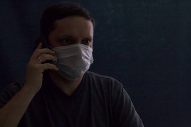 Tıbbi maskeli bir adam akıllı telefondan konuşuyor. Alacakaranlıkta. Koronavirüs sırasında kendini izole eden enfeksiyonlara karşı koruma.
