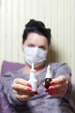 Tıbbi maskeli bir kadın yatakta oturuyor ve elinde burun spreyi kavanozları tutuyor. Koronavirüs sırasında kendini izole eden enfeksiyonlara karşı koruma.
