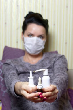 Tıbbi maskeli bir kadın yatakta oturuyor ve elinde burun spreyi kavanozları tutuyor. Koronavirüs sırasında kendini izole eden enfeksiyonlara karşı koruma.