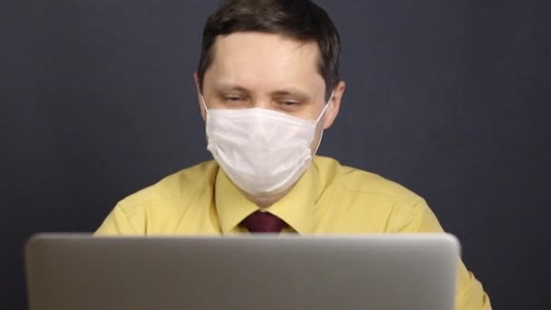 A man in a medical mask reads the news in a laptop. Laughs, squinting his eyes. Self-isolation during the coronavirus epidemic. — Stock Video