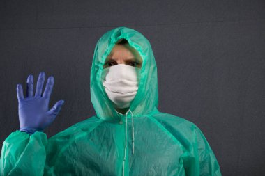 Koruyucu gazlı bez maskeli bir adam ve kapüşonlu bir yağmurluk. Lastik eldivenlerle. Sağ elinin avucuyla selam veriyor. Virüs enfeksiyonuna karşı basit bir koruma. Gri bir arka planda..
