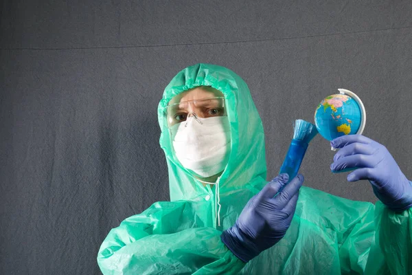 A man in a protective gauze mask, glasses and a raincoat with a hood. Holds a globe in his hand and brushes it with a brush. Hands in rubber gloves. Symbol of the global epidemic. It is on a gray background.