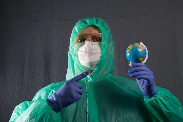 A man in a protective gauze mask, glasses and a raincoat with a hood. Holds a globe in front of him, and points a finger at it. Hands in rubber gloves. Symbol of the global epidemic. It is on a gray background.