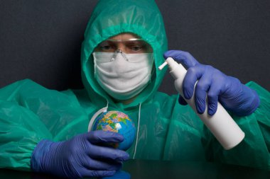 Koruyucu gazlı bez maskeli, gözlüklü ve kapüşonlu bir adam. Küreyi elinde tutuyor ve onu atomdan işliyor. Eldivenli eller. Küresel salgının sembolü. Gri bir arka planda..