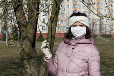 Koruyucu gazlı bez maskeli bir kadın söğüt ağacının yanında duruyor. İlkbaharda Avrupa 'daki salgın. Küresel salgının sembolü. Gri bir arka planda..