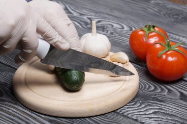 Lastik eldivenli bir adam kesme tahtasındaki bir bıçakla salatalığı kesiyor. Yakınlarda yeşil at kuyruklu ve sarımsaklı kırmızı domatesler var. Fırçalanmış çam ağaçlarının yüzeyinde.
