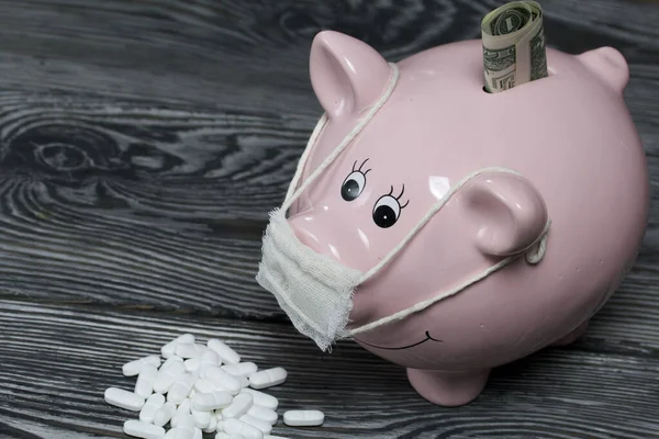 Ceramic piggy bank in pink. With a gauze bandage. A bill sticks out of it. In front of her is a handful of white pills. Treatment savings during an epidemic.