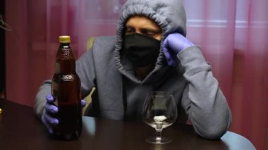 Koruyucu maske ve lastik eldiven takmış genç bir adam bir masada oturuyor, başını ellerinin üzerine koyuyor. Önünde bir şişe bira ve bir bardak var. Elden ele geziyor ve nasıl içileceğini düşünüyor. Ben...
