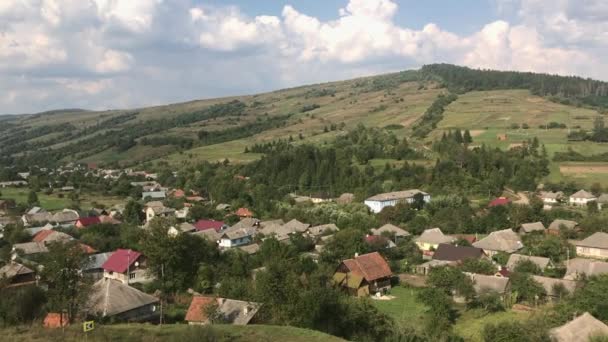 Un pueblo en un valle montañoso. Vista panorámica desde la colina. Las casas se encuentran abajo. Cielo azul, colinas verdes . — Vídeos de Stock