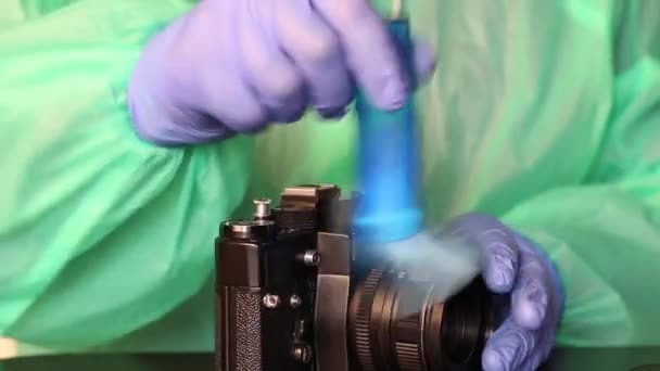 Um homem de luvas de borracha, uma capa de chuva e óculos remove poeira de uma câmera com um pincel. Limpeza de equipamentos fotográficos durante uma epidemia — Vídeo de Stock