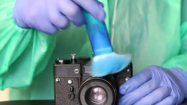 Een man met rubberen handschoenen, een regenjas en een bril verwijdert stof van een camera met een borstel. Schoonmaken van fotografische apparatuur tijdens een epidemie. — Stockvideo