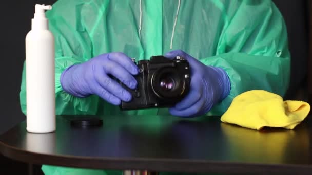 Un fotógrafo con guantes de goma y un impermeable aplica un antiséptico de una botella de spray a una servilleta. Se frota la cámara. Trabajo durante una pandemia . — Vídeo de stock