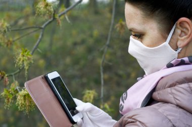 Bir kız, bir bahar parkında akıllı telefondan fotoğraf çekiyor. Yüzünde bir kumaş koruyucu maske var. Bahar ceketi giymişti, boynunda bir atkı vardı. Salgın sırasında temiz havada bir yürüyüş..