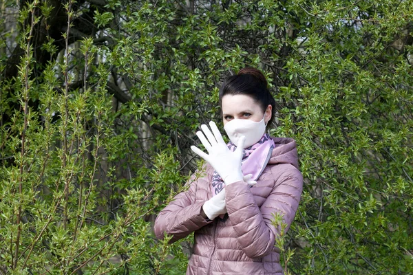 Portrait of a girl in a spring park. On the face a cloth protective mask, puts on gloves. Dressed in a spring jacket, a scarf around her neck. A walk in the fresh air during a pandemic.