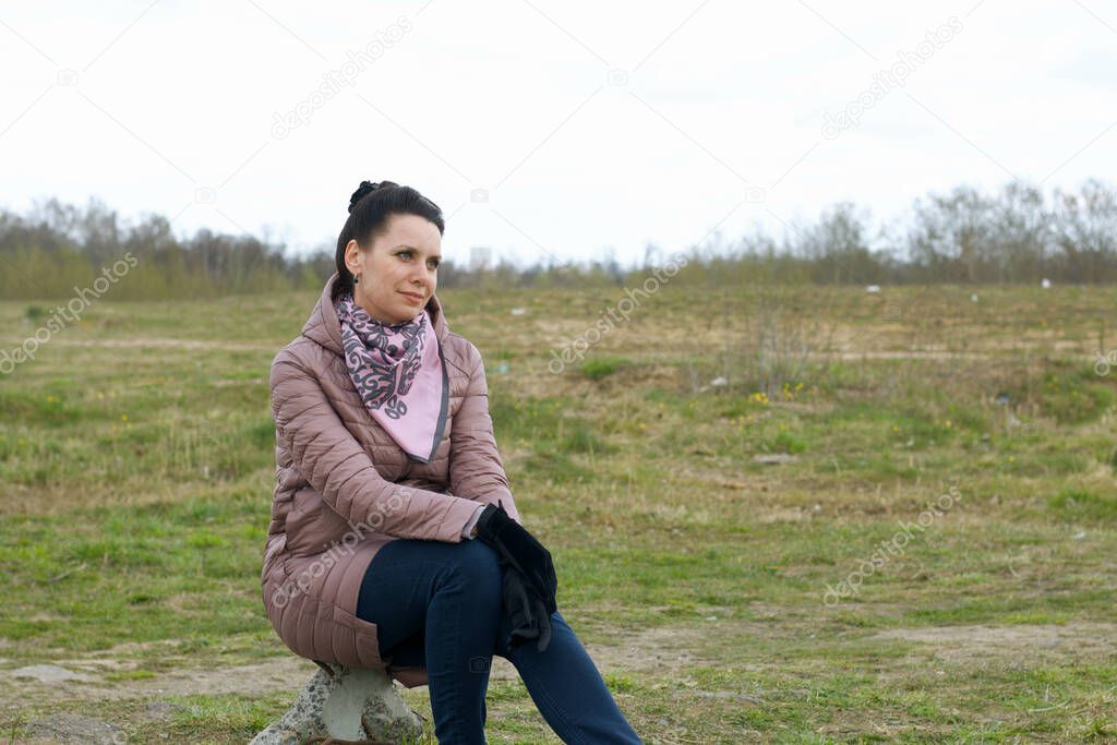 Girl in the spring meadow. Sits on a stone. Dressed in a spring jacket, a scarf around her neck.